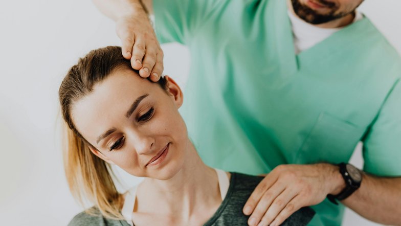 Eine Frau erhält eine Nackenmassage von einem männlichen Therapeuten. Der Therapeut hat seine Hand auf ihren Kopf gelegt, während sie entspannt aussieht. Beide tragen bequeme Kleidung.