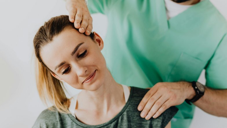 Eine Frau erhält eine Nackenmassage von einem männlichen Therapeuten. Der Therapeut hat seine Hand auf ihren Kopf gelegt, während sie entspannt aussieht. Beide tragen bequeme Kleidung.