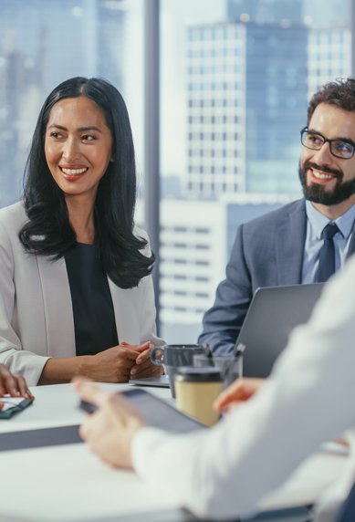 A group of five people in formal attire is sitting at a table in a modern office, engaging in discussion.