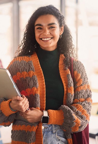 A smiling woman with curly hair stands in a modern learning space, holding a laptop and wearing a striped cardigan. In the background, other students can be seen working at tables.
