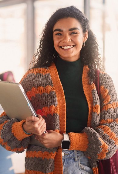 A smiling woman with curly hair stands in a modern learning space, holding a laptop and wearing a striped cardigan. In the background, other students can be seen working at tables.