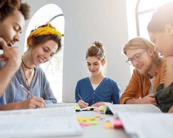 Five people are sitting at a table, collaborating on notes and documents while supporting each other and exchanging ideas.