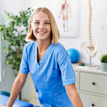 Eine lächelnde Frau in blauer Arbeitskleidung steht an einem Tisch in einem Behandlungsraum, umgeben von medizinischen Geräten und Pflanzen.