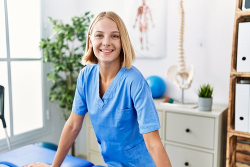 Eine lächelnde Frau in blauer Arbeitskleidung steht an einem Tisch in einem Behandlungsraum, umgeben von medizinischen Geräten und Pflanzen.