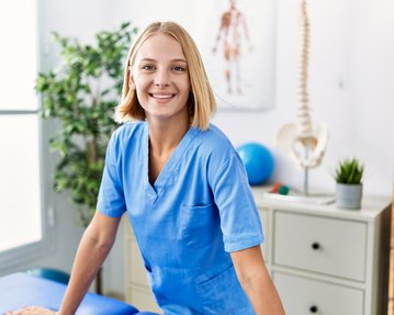 Eine lächelnde Frau in blauer Arbeitskleidung steht an einem Tisch in einem Behandlungsraum, umgeben von medizinischen Geräten und Pflanzen.
