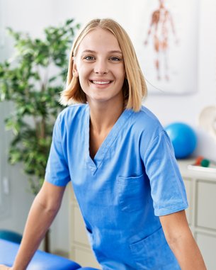 Eine lächelnde Frau in blauer Arbeitskleidung steht an einem Tisch in einem Behandlungsraum, umgeben von medizinischen Geräten und Pflanzen.