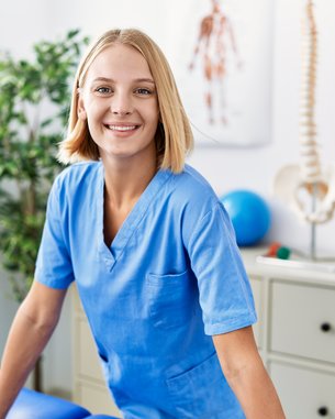Eine lächelnde Frau in blauer Arbeitskleidung steht an einem Tisch in einem Behandlungsraum, umgeben von medizinischen Geräten und Pflanzen.