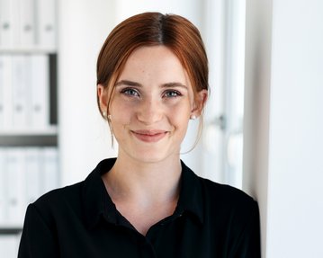Junge Frau mit roten Haaren, die in einem Büro steht und lächelt. Im Hintergrund sind Aktenordner sichtbar.