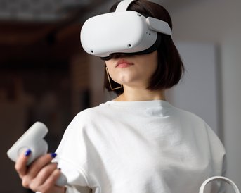 Eine Frau trägt ein VR-Headset und hält einen Controller in der Hand. Sie schaut konzentriert und scheint in eine virtuelle Welt einzutauchen. Der Hintergrund ist unscharf und modern gestaltet.