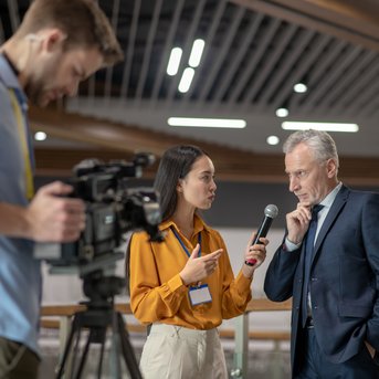 Eine Frau interviewt einen Mann mit grauen Haaren, während ein Kameramann im Hintergrund filmt. Beide tragen formelle Kleidung.