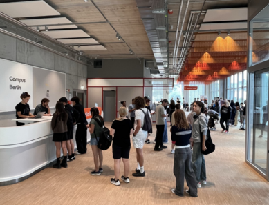 Menschen stehen an einem Empfangstresen in einem modernen Raum mit Holzfußboden. Im Hintergrund sind weitere Personen und eine große Fensterfront sichtbar. Über dem Empfang steht "Campus Berlin".