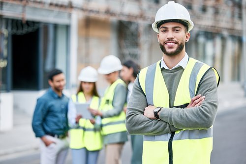 A construction manager in safety gear and a helmet stands smiling in the foreground, while a team of construction workers discusses in the background.
