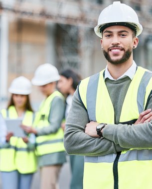 A construction manager in safety gear and a helmet stands smiling in the foreground, while a team of construction workers discusses in the background.