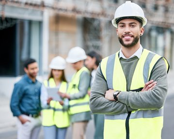 A construction manager in safety gear and a helmet stands smiling in the foreground, while a team of construction workers discusses in the background.