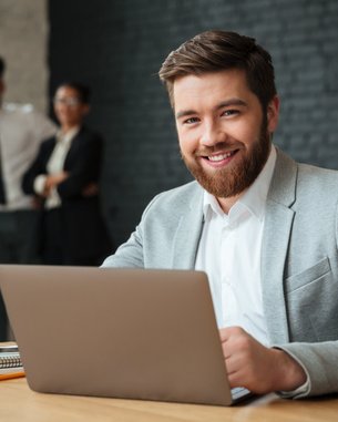 A man with a beard is sitting at a table, working on a laptop. He is wearing a gray suit and smiling. In the background, three people are standing in formal attire.