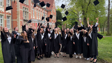 A group of graduates in black gowns throwing their caps in the air in front of a historic building outdoors.