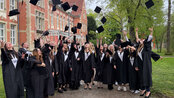 A group of graduates in black gowns throwing their caps in the air in front of a historic building outdoors.