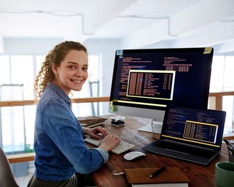 Eine junge Frau sitzt an einem Schreibtisch und arbeitet an zwei Computern, während sie lächelt. Auf den Bildschirmen sind Programmiercodes sichtbar.