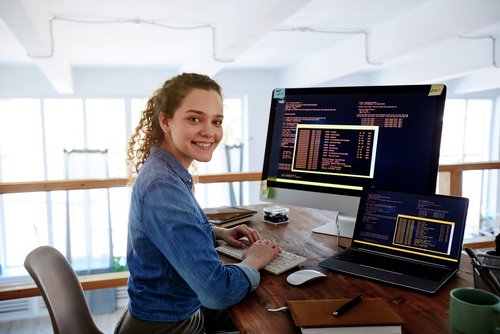 Eine junge Frau sitzt an einem Schreibtisch und arbeitet an zwei Computern, während sie lächelt. Auf den Bildschirmen sind Programmiercodes sichtbar.