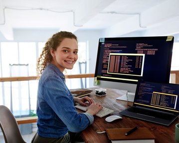 Eine junge Frau sitzt an einem Schreibtisch und arbeitet an zwei Computern, während sie lächelt. Auf den Bildschirmen sind Programmiercodes sichtbar.