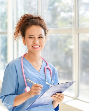 Eine junge Frau in blauer medizinischer Kleidung hält ein Clipboard und einen Stift. Sie trägt ein Stethoskop um den Hals und lächelt freundlich. Helle Fenster im Hintergrund lassen viel Licht herein.