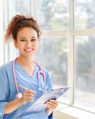 Eine junge Frau in blauer medizinischer Kleidung hält ein Clipboard und einen Stift. Sie trägt ein Stethoskop um den Hals und lächelt freundlich. Helle Fenster im Hintergrund lassen viel Licht herein.