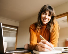 Eine lächelnde Frau in einem orangefarbenen Pullover schreibt in ein Notizbuch, während sie an einem Tisch mit einem Laptop sitzt.