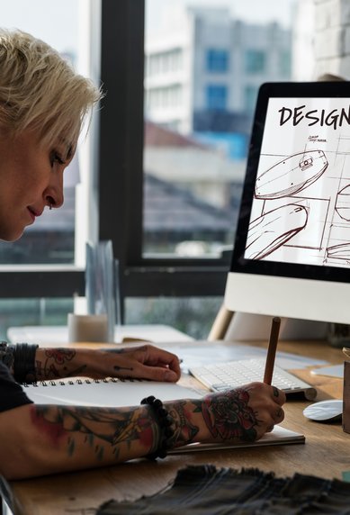 A person with blonde hair and tattoos sketches on a notepad while looking at a computer screen filled with designs.