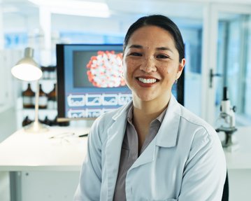Eine Wissenschaftlerin in einem Labor lächelt, trägt einen weißen Laborkittel und sitzt vor einem Computerbildschirm mit wissenschaftlichen Grafiken.