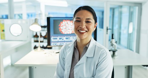 A scientist in a lab smiles, wearing a white lab coat, and sits in front of a computer screen displaying scientific graphs.