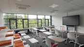 Moderne Klassenzimmer mit Tischen, Stühlen und einer großen Bildschirmwand. Orangefarbene Kissen auf einer erhöhten Sitzfläche.