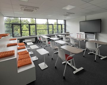 Modern classrooms with tables, chairs, and a large screen wall. Orange cushions on a raised seating area.