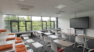 Moderne Klassenzimmer mit Tischen, Stühlen und einer großen Bildschirmwand. Orangefarbene Kissen auf einer erhöhten Sitzfläche.