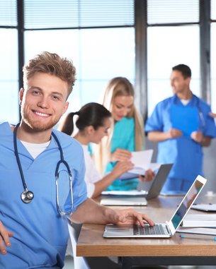 Ein junger Mann in blauer Arbeitskleidung mit Stethoskop sitzt an einem Tisch mit einem Laptop. Im Hintergrund sind mehrere Personen in medizinischer Kleidung, die miteinander sprechen und arbeiten.