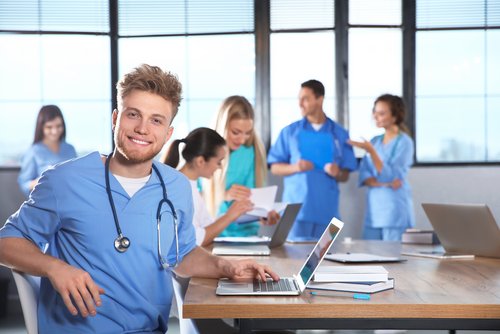 Ein junger Mann in blauer Arbeitskleidung mit Stethoskop sitzt an einem Tisch mit einem Laptop. Im Hintergrund sind mehrere Personen in medizinischer Kleidung, die miteinander sprechen und arbeiten.