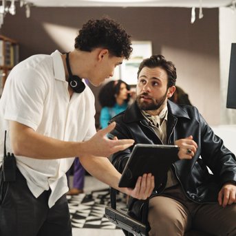 Zwei Männer in einem Studio diskutieren, während einer ein Tablet hält. Der andere trägt eine Lederjacke und hört aufmerksam zu.