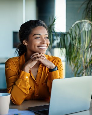 Eine Frau mit lockigem Haar sitzt an einem Tisch, lächelt und schaut auf einen Laptop. Vor ihr steht eine Tasse und einige Unterlagen liegen auf dem Tisch. Pflanzen sind im Hintergrund sichtbar.