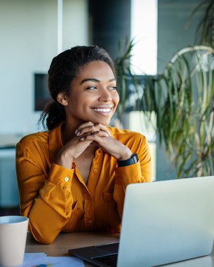 A woman with curly hair is sitting at a table, smiling and looking at a laptop. In front of her is a cup, and some documents are spread out on the table. Plants are visible in the background.