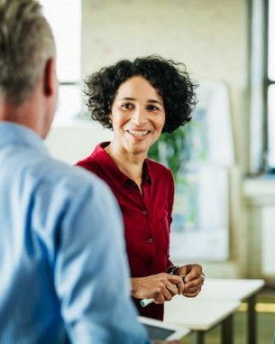 Eine Gruppe von vier Personen steht in einem modernen Büro und unterhält sich. Eine Frau mit lockigem Haar trägt ein rotes Hemd und lächelt, während die anderen Personen aufmerksam zuhören.