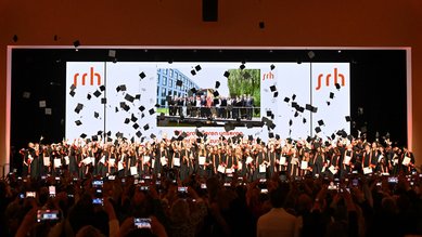 Graduates throw their caps in the air during a graduation ceremony, surrounded by cheering spectators and cameras.