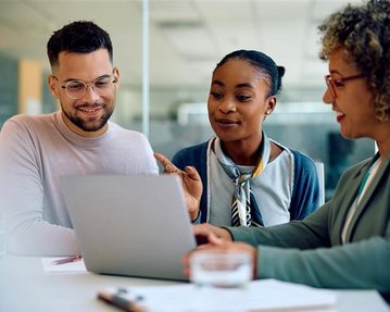 Drei Personen sitzen an einem Tisch und schauen auf einen Laptop. Sie diskutieren und zeigen auf den Bildschirm.
