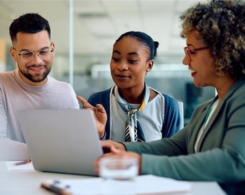 Three people are sitting at a table, looking at a laptop. They are discussing and pointing at the screen.