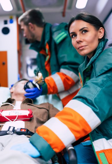 Zwei Sanitäter in einem Krankenwagen kümmern sich um einen Patienten, der auf einer Trage liegt. Medizingeräte sind sichtbar.