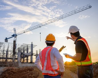 Two construction workers in safety vests and helmets are looking at a construction project with a crane in the background as the sun sets.