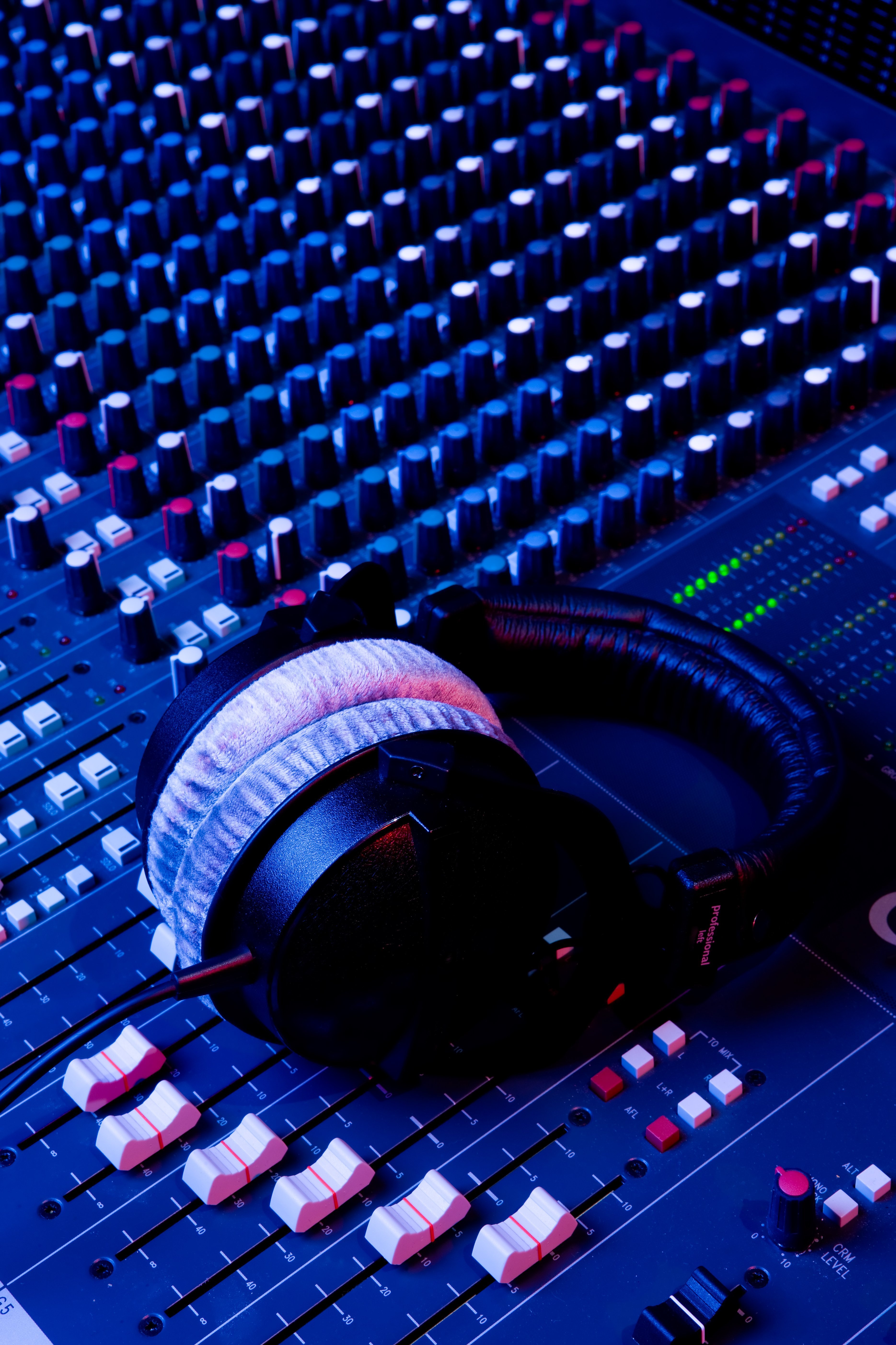 Headphones rest on a mixing console filled with numerous knobs and switches. The surface of the console is illuminated, and the knobs are arranged in various colors. The scene conveys a professional audio production environment.