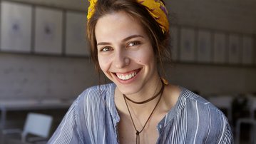 Eine lächelnde Frau mit einem gelben Haarband sitzt in einem modernen Café. Sie trägt ein gestreiftes Oberteil und hat eine freundliche Ausstrahlung. Im Hintergrund sind verschwommene Möbel zu sehen.