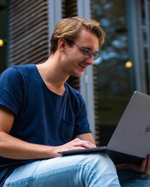 Ein junger Mann sitzt draußen und arbeitet an einem Laptop. Er trägt ein blaues T-Shirt und eine Brille. Der Hintergrund zeigt Holzlamellen und unscharfe Bäume.