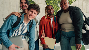 A group of four young people smiles happily as they stand together, holding laptops and documents. In the background, there is a green world map made of plants.