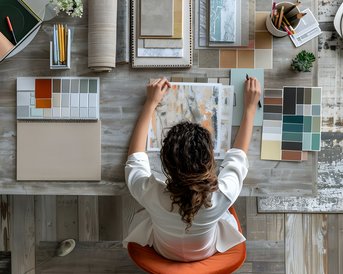 A person is sitting at a table, surrounded by various color patterns and materials, while working on a creative project. The table is neat and well-organized.