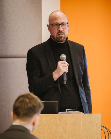 Ein Professor steht an einem Podium, hält ein Mikrofon und spricht zu einem Publikum. Im Hintergrund ist ein Bildschirm mit Text.